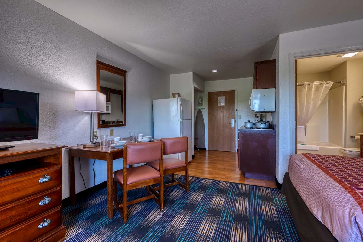 The image shows a hotel room with a bed, a table with chairs, a TV, a fridge, and a bathroom. The decor is simple and functional.